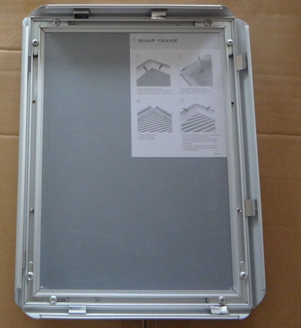 A product shot of the back of an open aluminium snap frame, showing the hinges, secure backing board, and an instruction diagram on how to use the frame's clip mechanism.