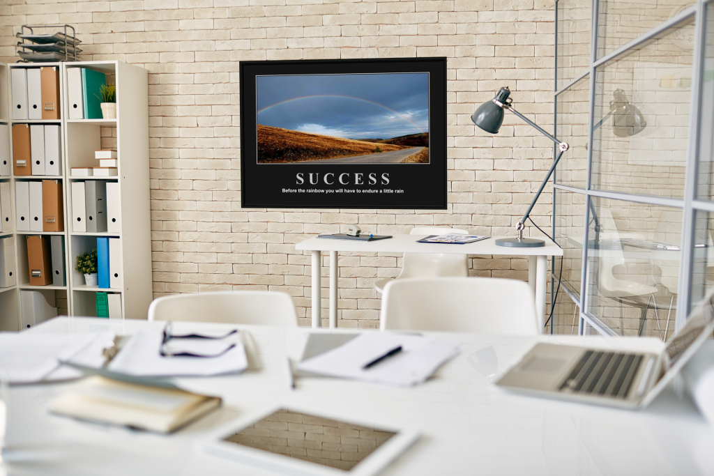A "Success" motivational poster with a rainbow over a landscape, displayed as a framed motivational poster in a modern, well-lit office workspace.